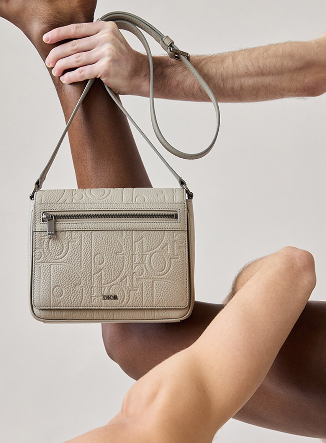Tan 'Dior' embossed leather messenger bag with adjustable shoulder strap, showcased between dark and light-skinned arm.