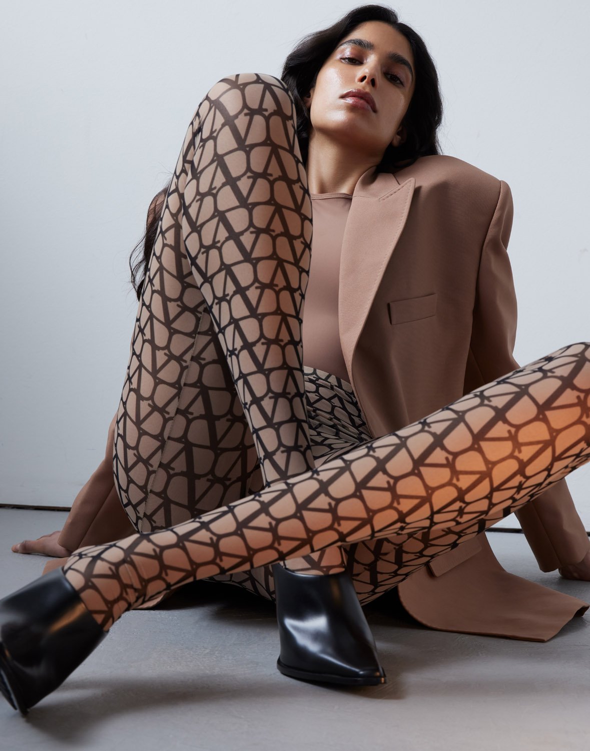 Model seated wearing printed patterned tights and a tan blazer against a grey studio floor background