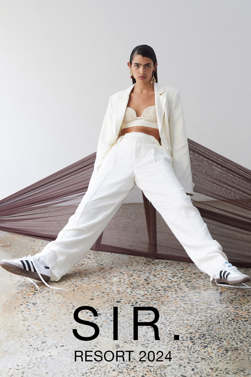 Woman poses in a white blazer, crop top, and wide-leg pants, with Adidas sneakers, in a fashion editorial with a draped backdrop.