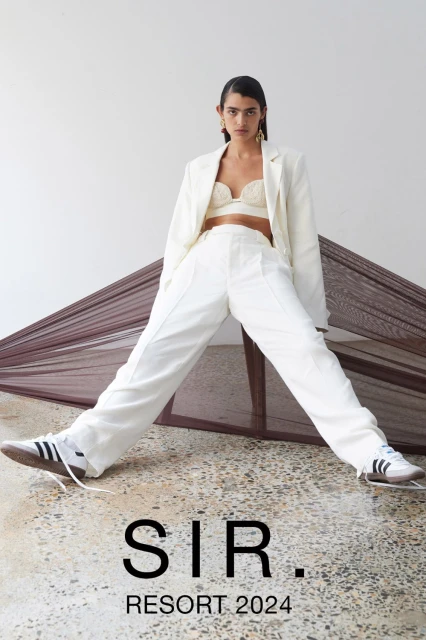 Woman poses in a white blazer, crop top, and wide-leg pants, with Adidas sneakers, in a fashion editorial with a draped backdrop.