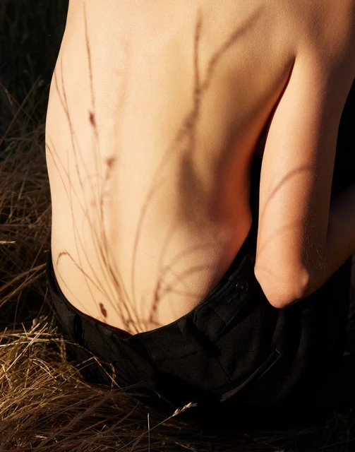 Close-up of a person's back with grass shadows, sitting in dried grass wearing black pants, illuminated by warm sunlight.