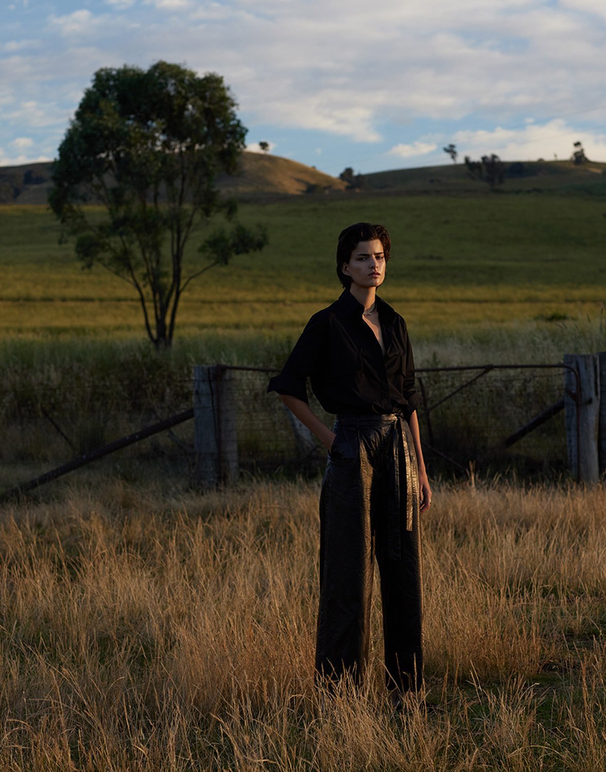 A woman with short hair stands in a grassy field, wearing a black shirt and dark pants, with a tree and rolling hills in the background.