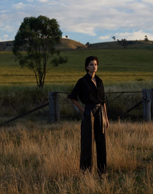 A woman with short hair stands in a grassy field, wearing a black shirt and dark pants, with a tree and rolling hills in the background.