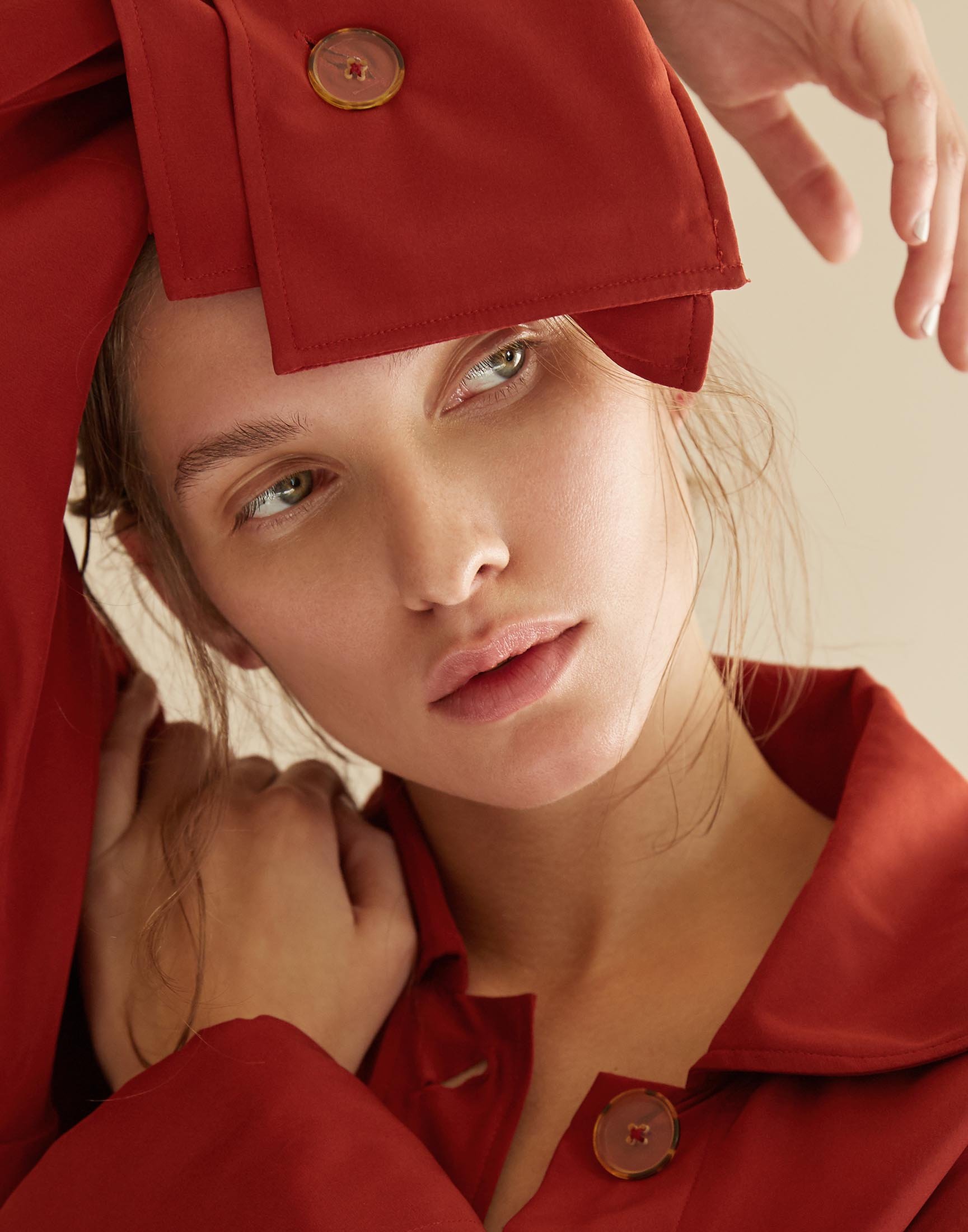 Close-up portrait of a young adult woman with fair skin wearing a red coat, posing with hands near her face, against a neutral backdrop.
