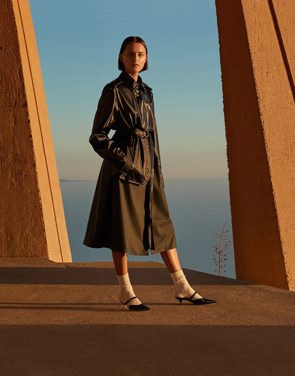 Fashion shot of a woman in a belted olive-green coat, white socks, and kitten heels, posing against a tan-colored building with a view of the sea.
