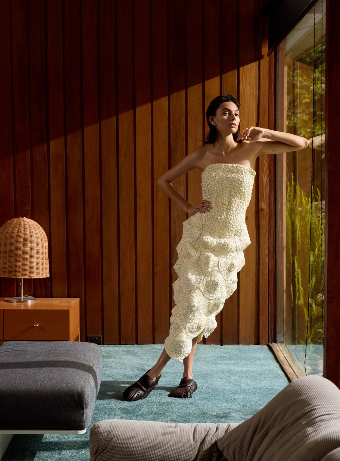 Elegant woman in textured strapless gown leans on glass wall in modern room with wood wall, side table and lamp, on blue carpet.