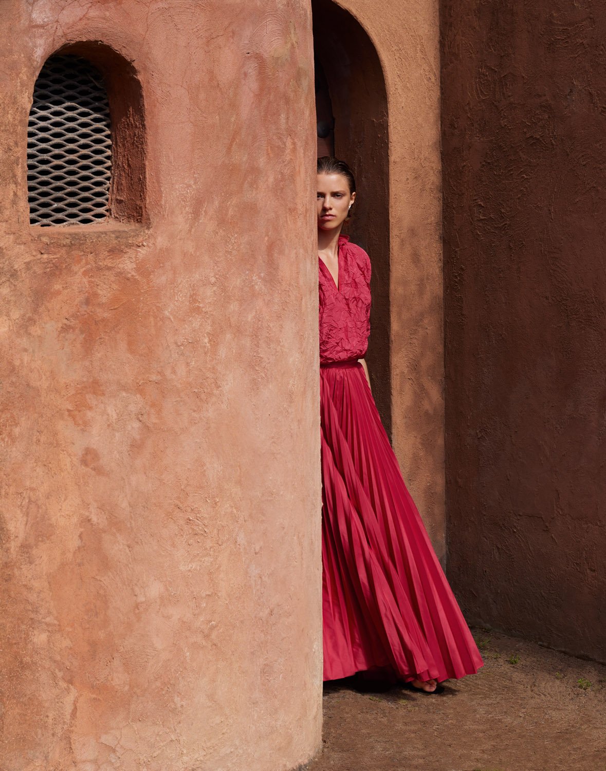 Model in vibrant red pleated skirt and top peeking from behind a curved terracotta architectural archway.