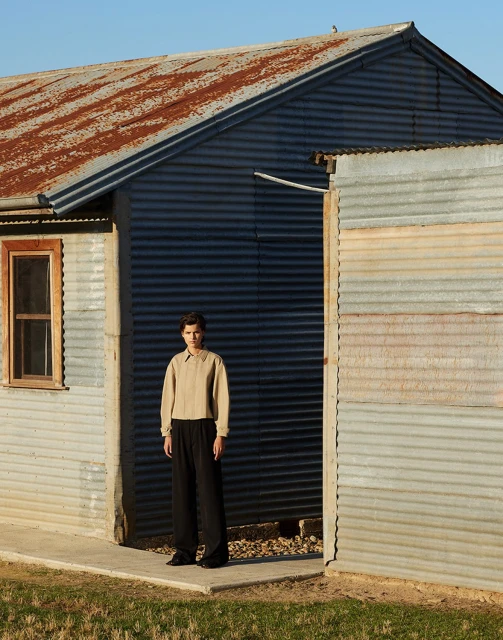 Full body shot of a young adult in a tan shirt and black pants, standing in front of a corrugated metal building during daytime.