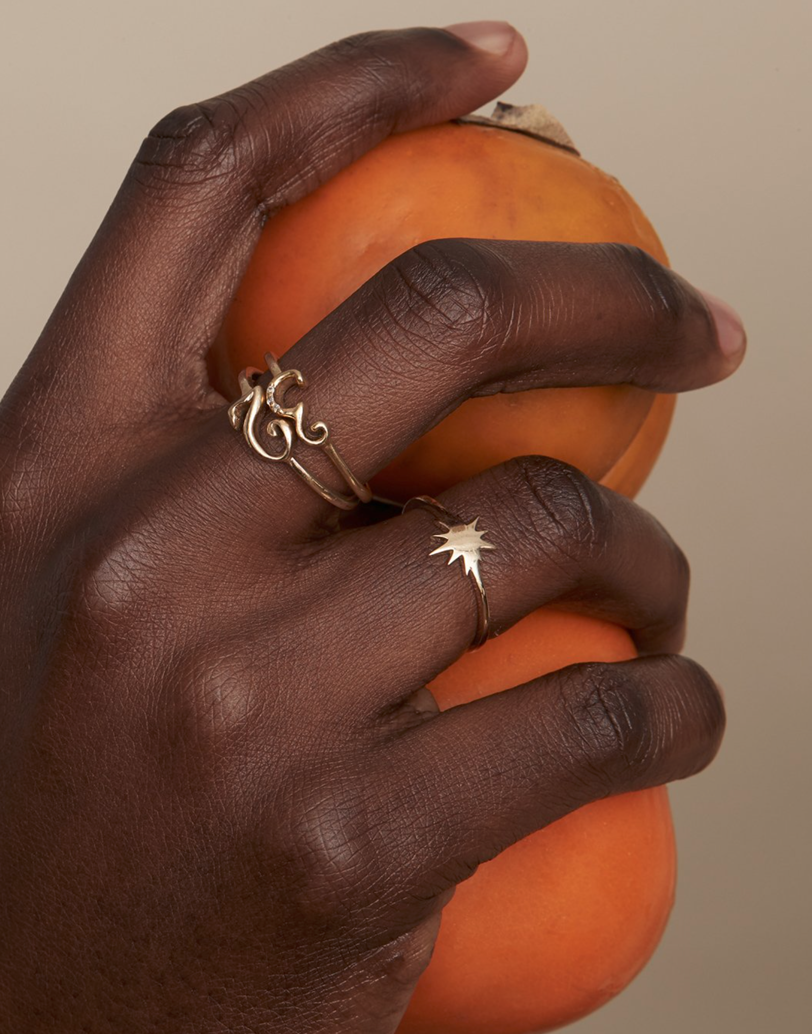 Close-up of a dark-skinned hand holding two persimmons, adorned with golden rings, set against a neutral, soft background.