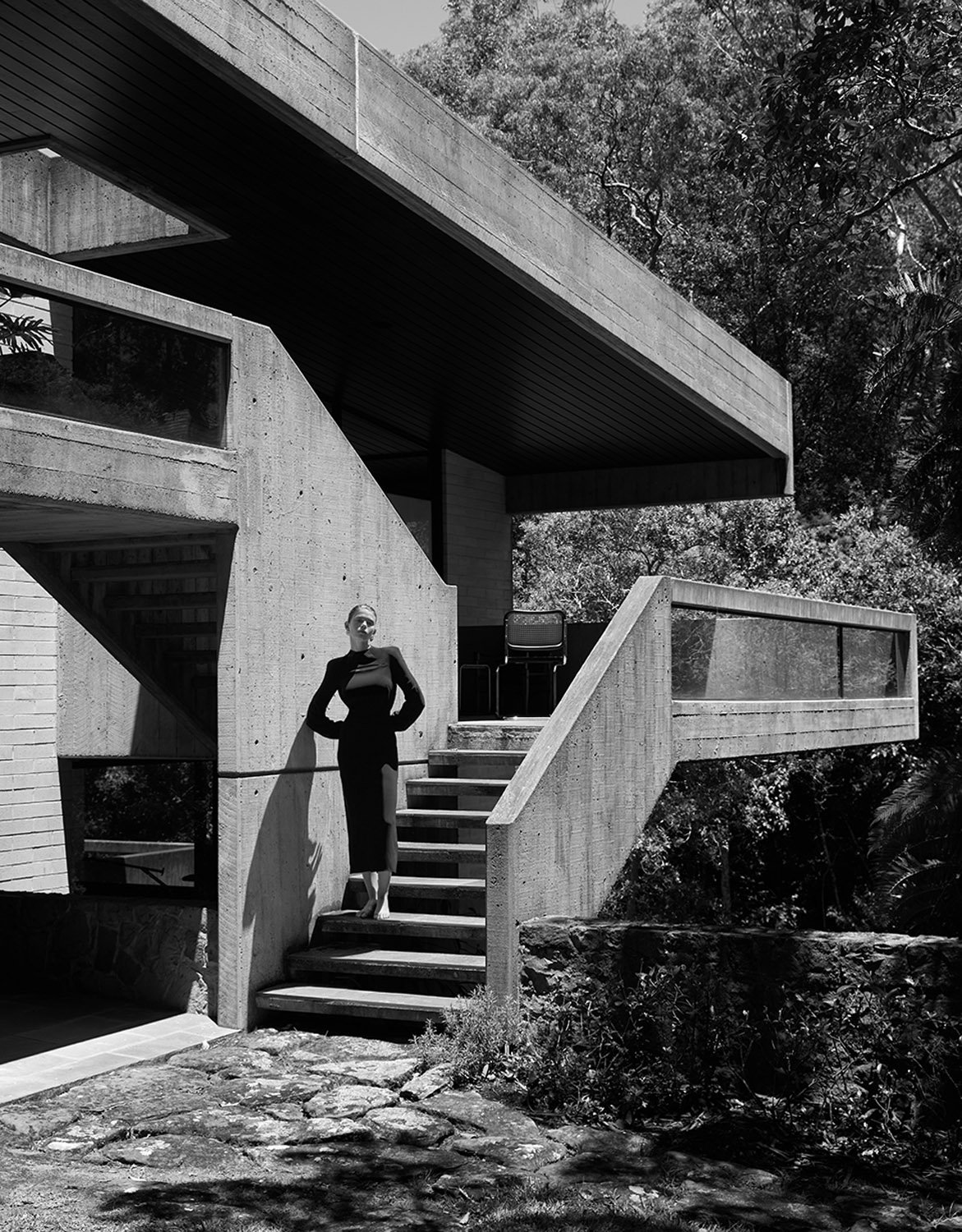 Black and white shot of a woman in a dress posing on the concrete steps of a brutalist-style house surrounded by trees.