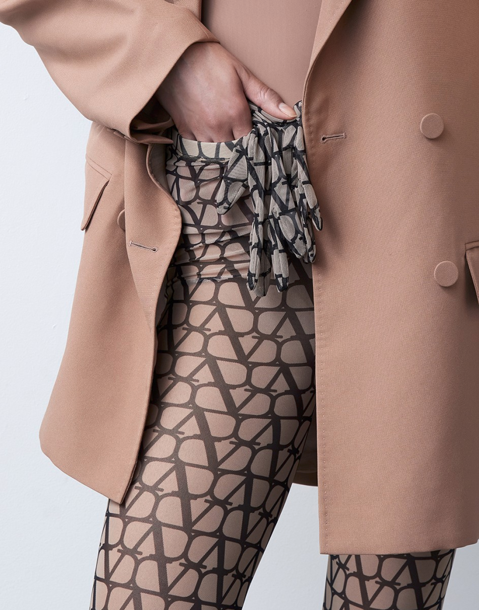 Close-up of a woman wearing patterned tights and a beige blazer with a beige top and patterned gloves in the pocket.