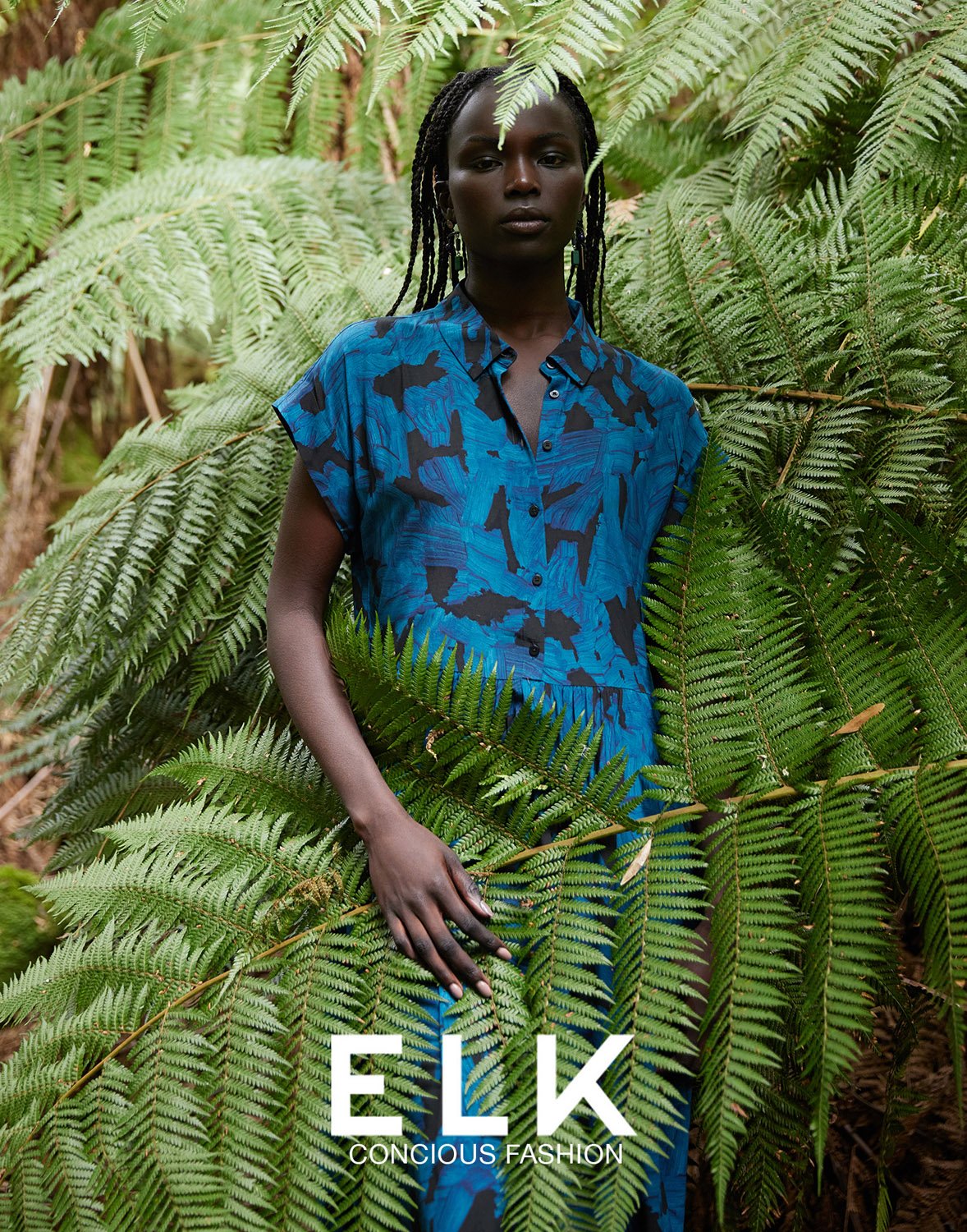 Fashion portrait of Black female model in blue printed dress amidst green ferns, promoting Elk Conscious Fashion brand outdoors.
