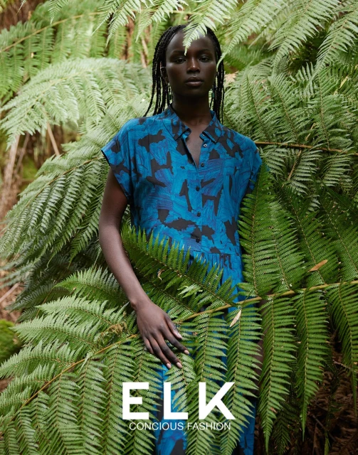 Fashion portrait of Black female model in blue printed dress amidst green ferns, promoting Elk Conscious Fashion brand outdoors.