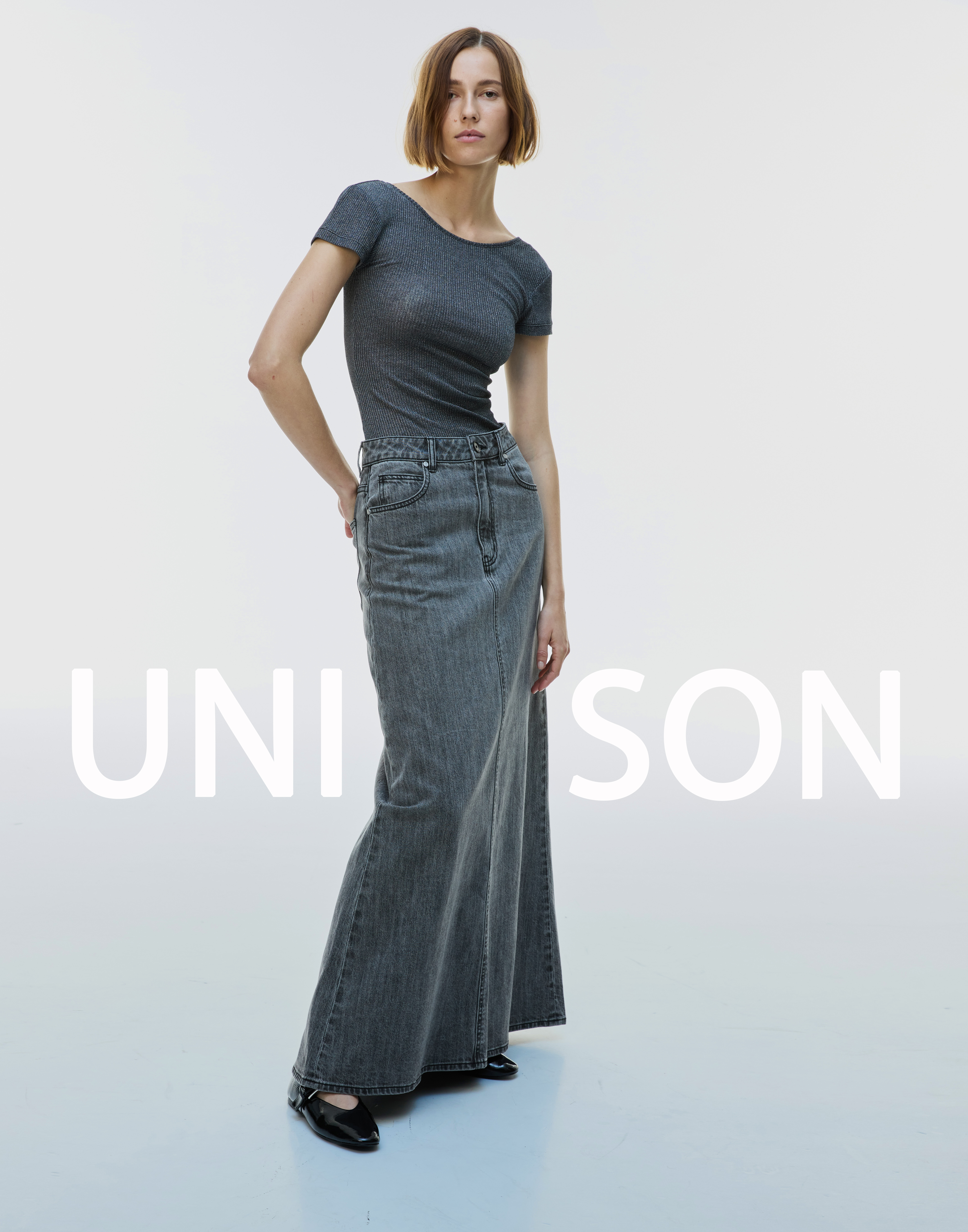 A fair-skinned woman models gray casual top and denim maxi skirt in a bright studio with soft lighting, fashion editorial.