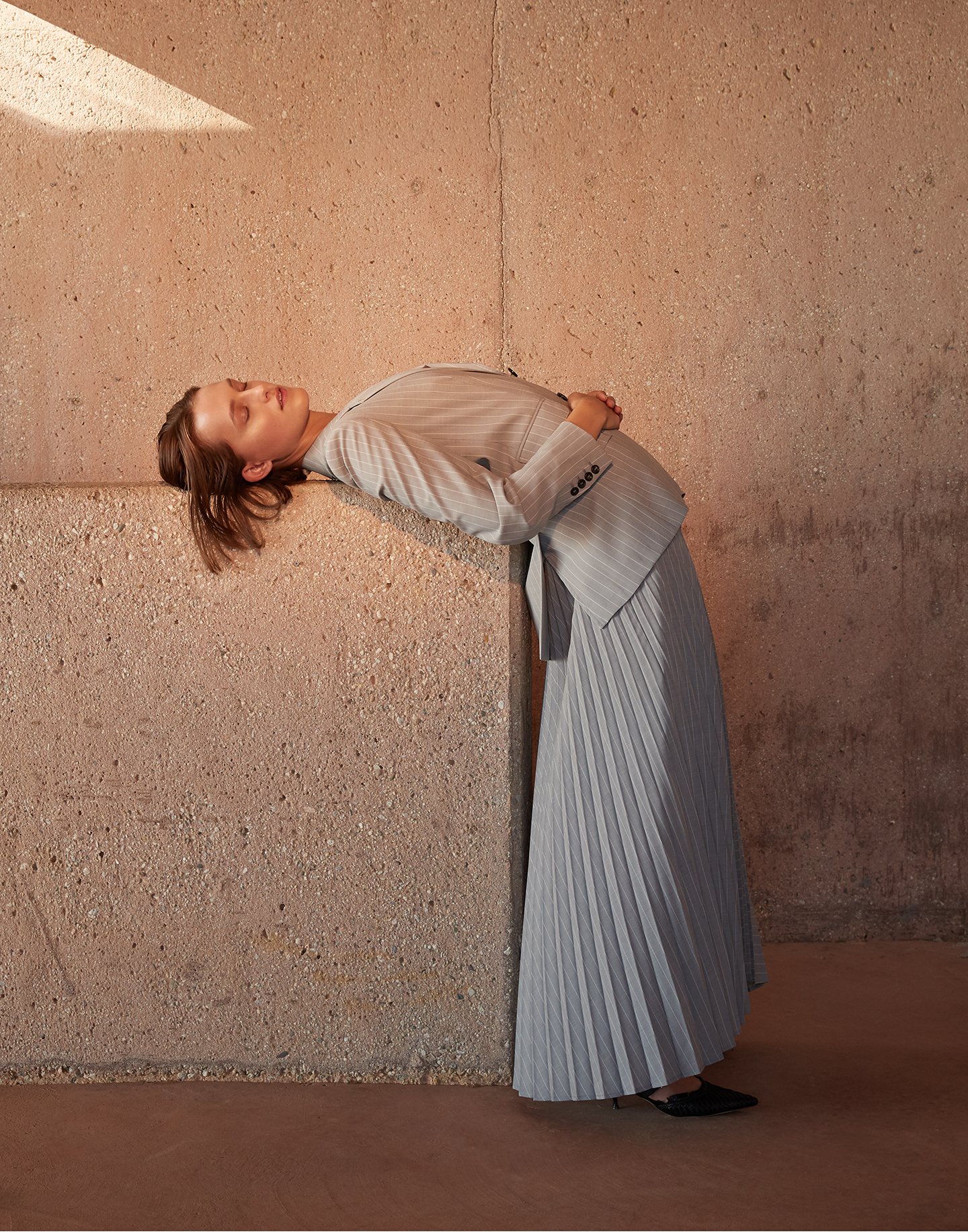 Woman in a grey skirt suit leaning against a concrete wall bathed in sunlight, creating a minimalist and elegant fashion portrait.