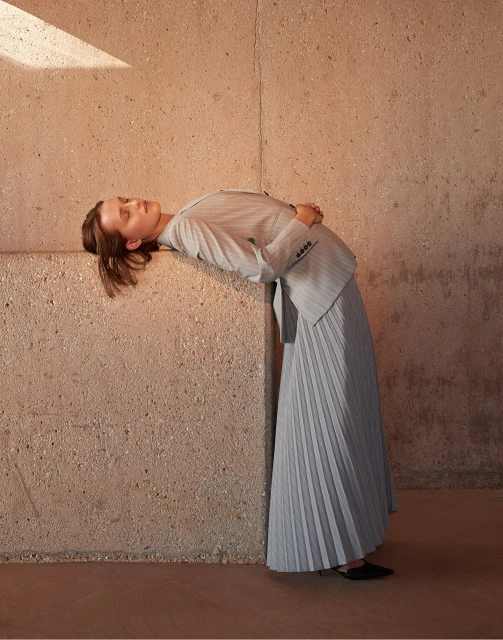 Woman in a grey skirt suit leaning against a concrete wall bathed in sunlight, creating a minimalist and elegant fashion portrait.