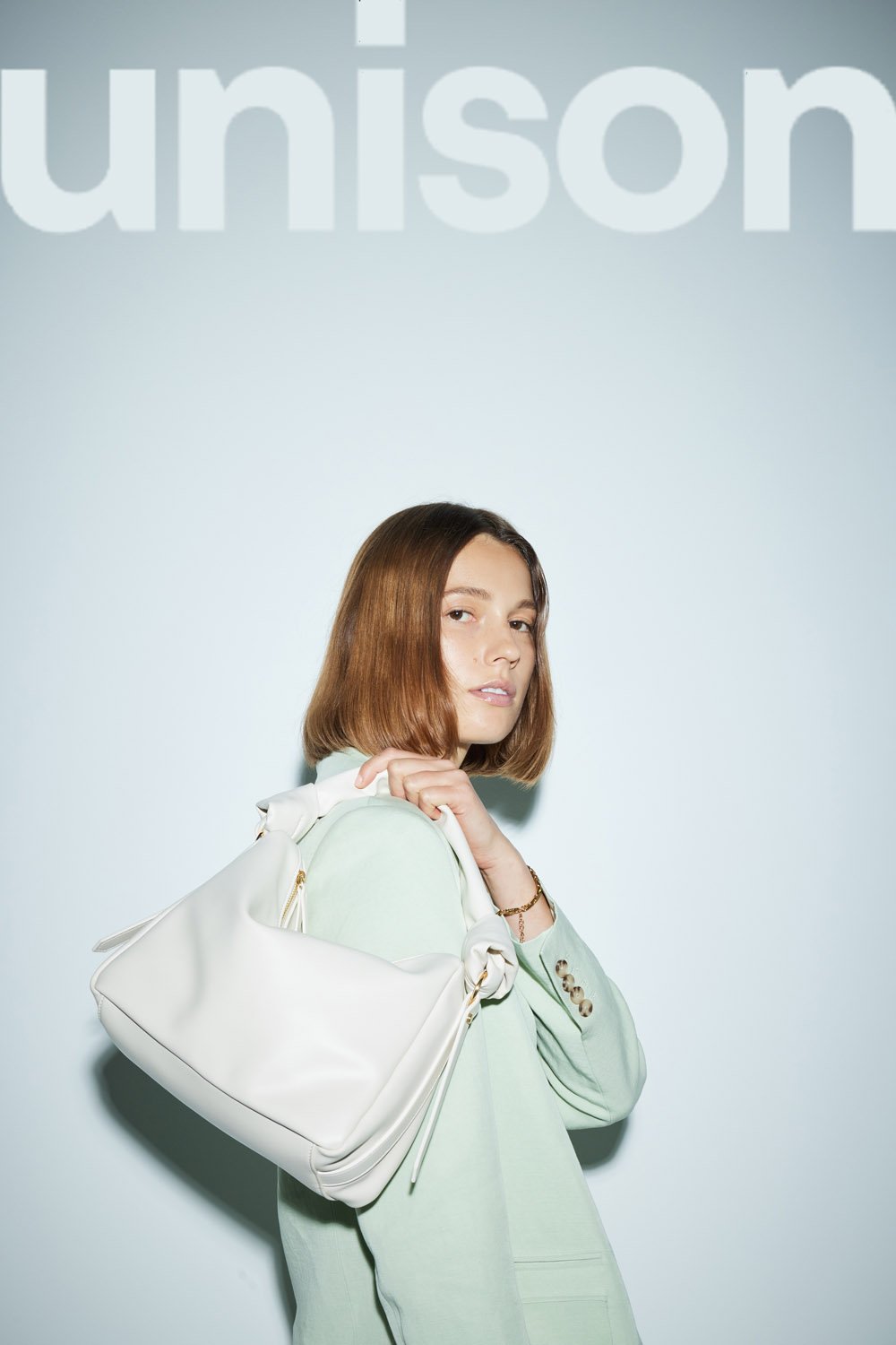 An adult woman poses indoors with a cream-colored handbag over her shoulder, wearing a mint-green jacket against a light-blue backdrop.