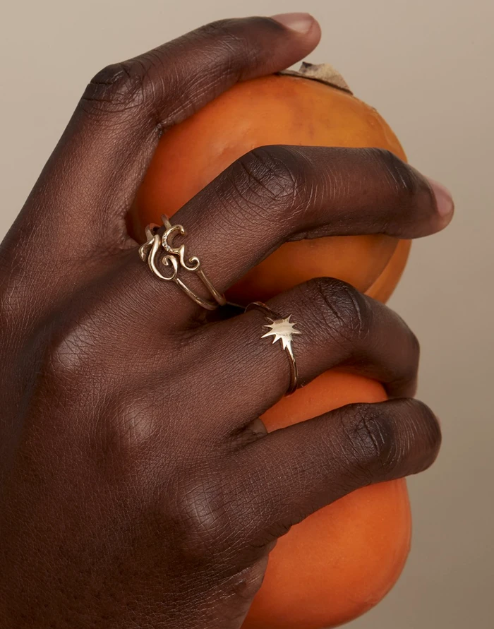 Close-up of hands holding two round orange fruits wearing gold rings against a neutral backdrop