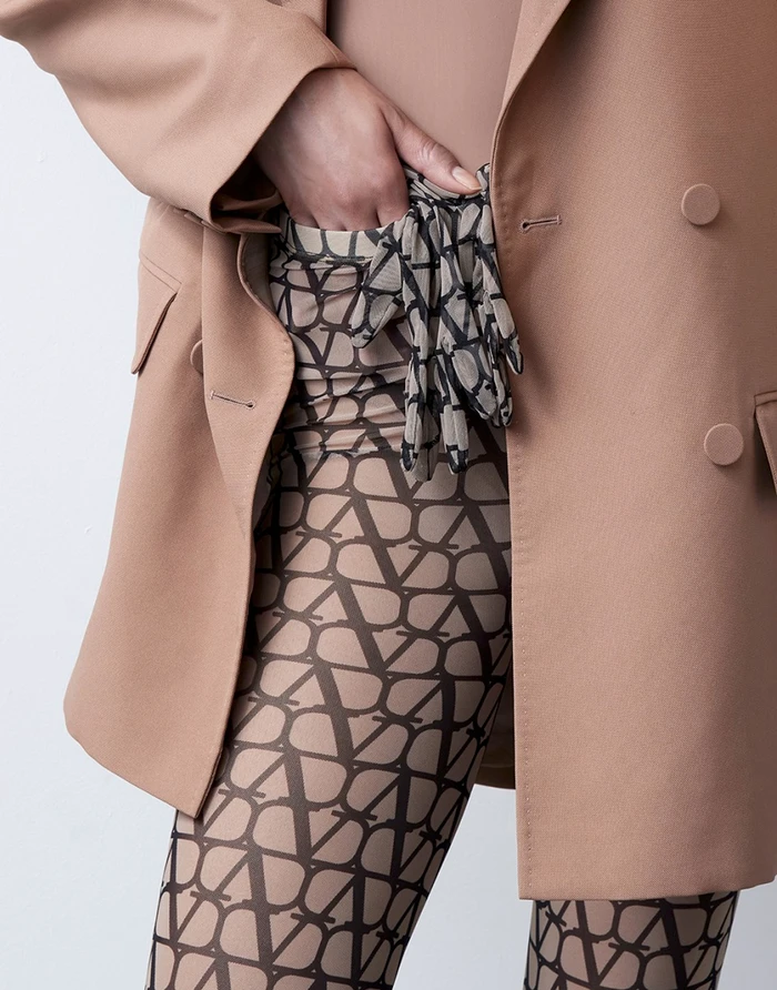 Close-up of a person wearing a beige blazer over monogram patterned tights with a hand tucked into the pocket