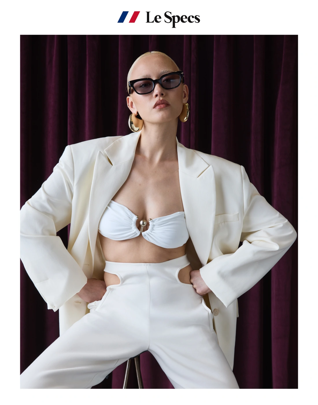 Model with shaved head wearing dark sunglasses, white blazer, white crop top, and white pants against a deep purple curtain background