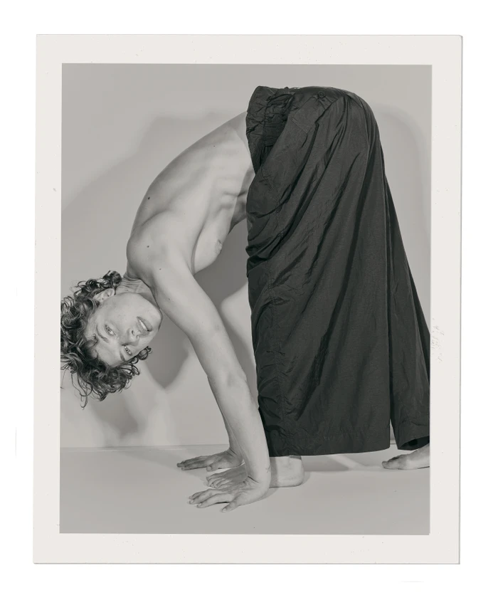 Man with curly hair bending over backwards wearing black trousers on a white studio background