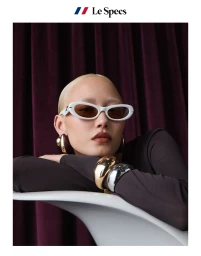 Model with bleached blonde hair wearing white sunglasses, dark long-sleeve top, and gold and silver bracelets leaning on a white curved rail against a dark curtain background