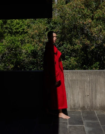 Model wearing a long red coat standing on a stone terrace with lush green foliage in the background.