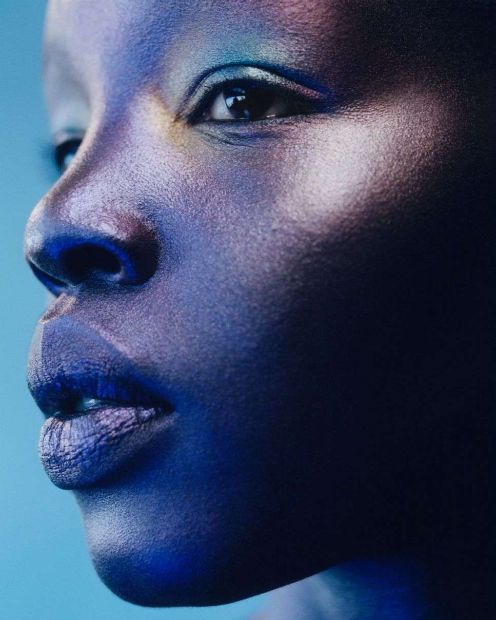 Close-up portrait of a Black woman with iridescent blue makeup on her face against a blue background