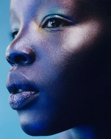 Close-up portrait of a Black woman with iridescent blue makeup on her face against a blue background