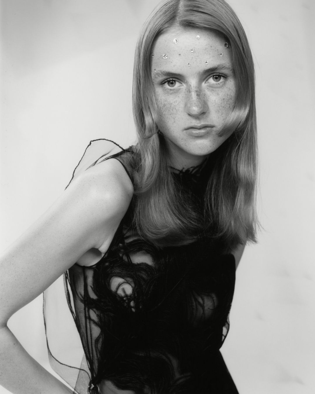Portrait of a young fair-skinned woman with long blonde hair wearing a black sleeveless top with decorative embellishments against a light background.