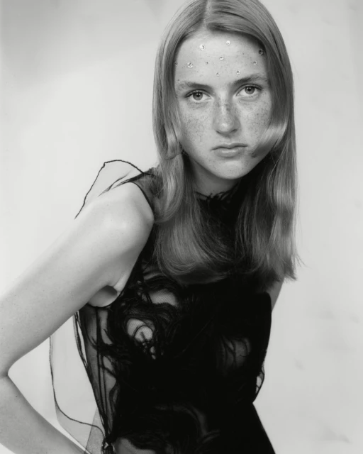 Portrait of a young fair-skinned woman with long blonde hair wearing a black sleeveless top with decorative embellishments against a light background.