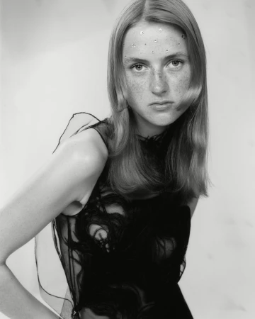 Portrait of a young fair-skinned woman with long blonde hair wearing a black sleeveless top with decorative embellishments against a light background.