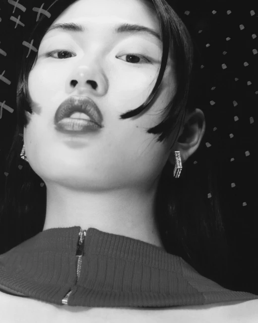 Monochrome portrait of an Asian female with bold lipstick, wearing earrings, and a red top against a dark patterned backdrop.