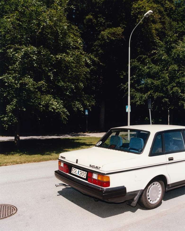 White Volvo 244 sedan parked on an asphalt road with green trees and a streetlight in the background on a sunny day.