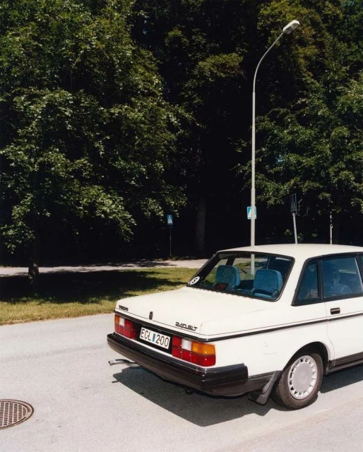 White Volvo 244 sedan parked on an asphalt road with green trees and a streetlight in the background on a sunny day.