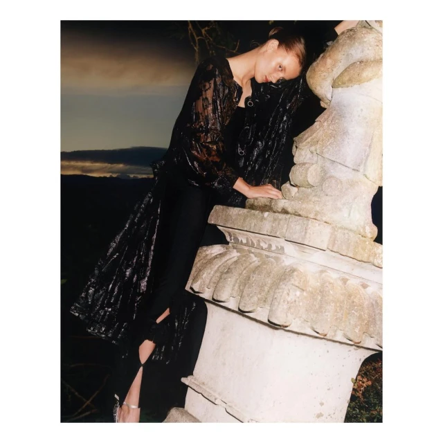 Fashionable woman in a sheer top leans against a stone sculpture at dusk. Mountains are visible in the background.