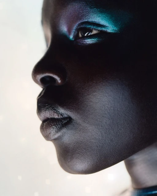Close-up portrait of a Black woman in profile with dramatic blue and purple makeup on her eyelids and cheekbones against a bright background.