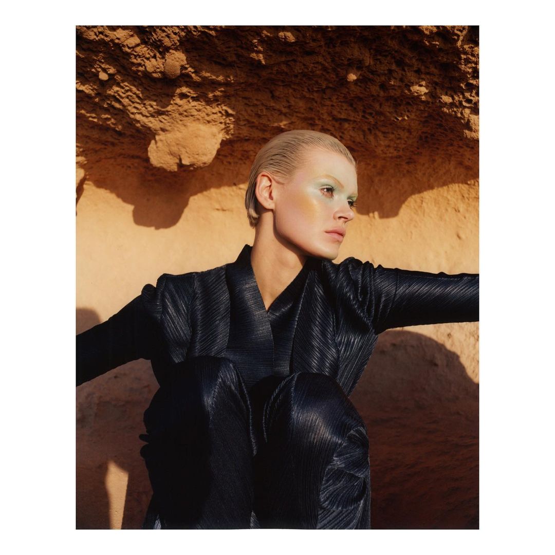 Fashion shot of a woman in black shiny outfit with slicked back hair, wearing green eyeshadow, sitting in front of a textured brown wall.