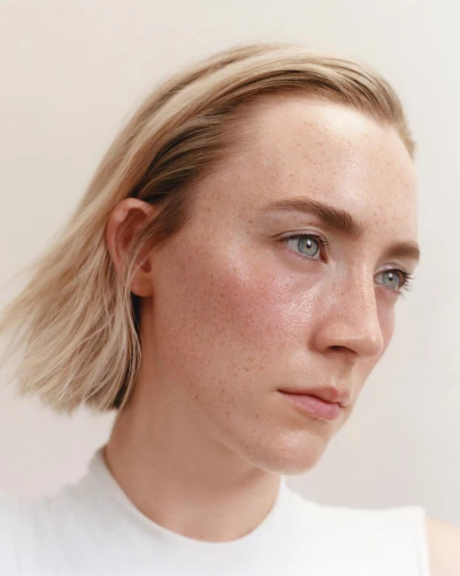 Portrait of a fair-skinned woman with blonde hair and freckles. She is wearing a white top, and the background is a solid neutral color.