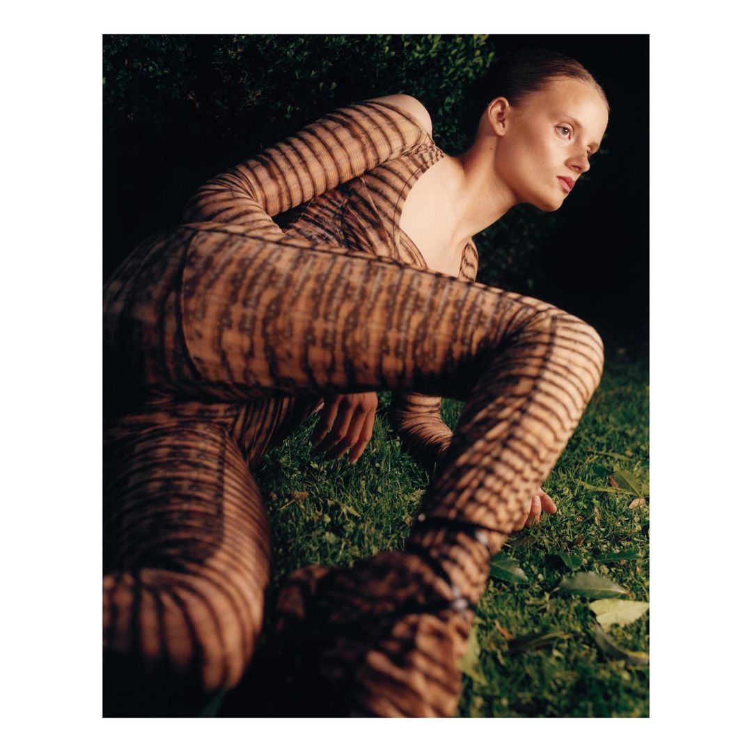 Adult female model in striped bodysuit crouching in a grassy outdoor setting with dark green foliage in the background, fashion editorial.