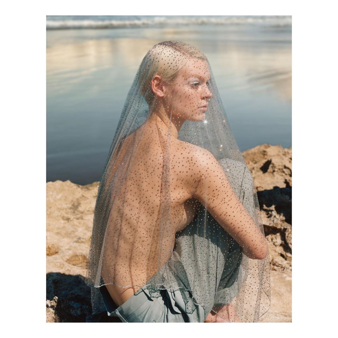 Young adult woman with short blonde hair and draped in shimmering net fabric poses on a rocky outcrop against a blue ocean backdrop.