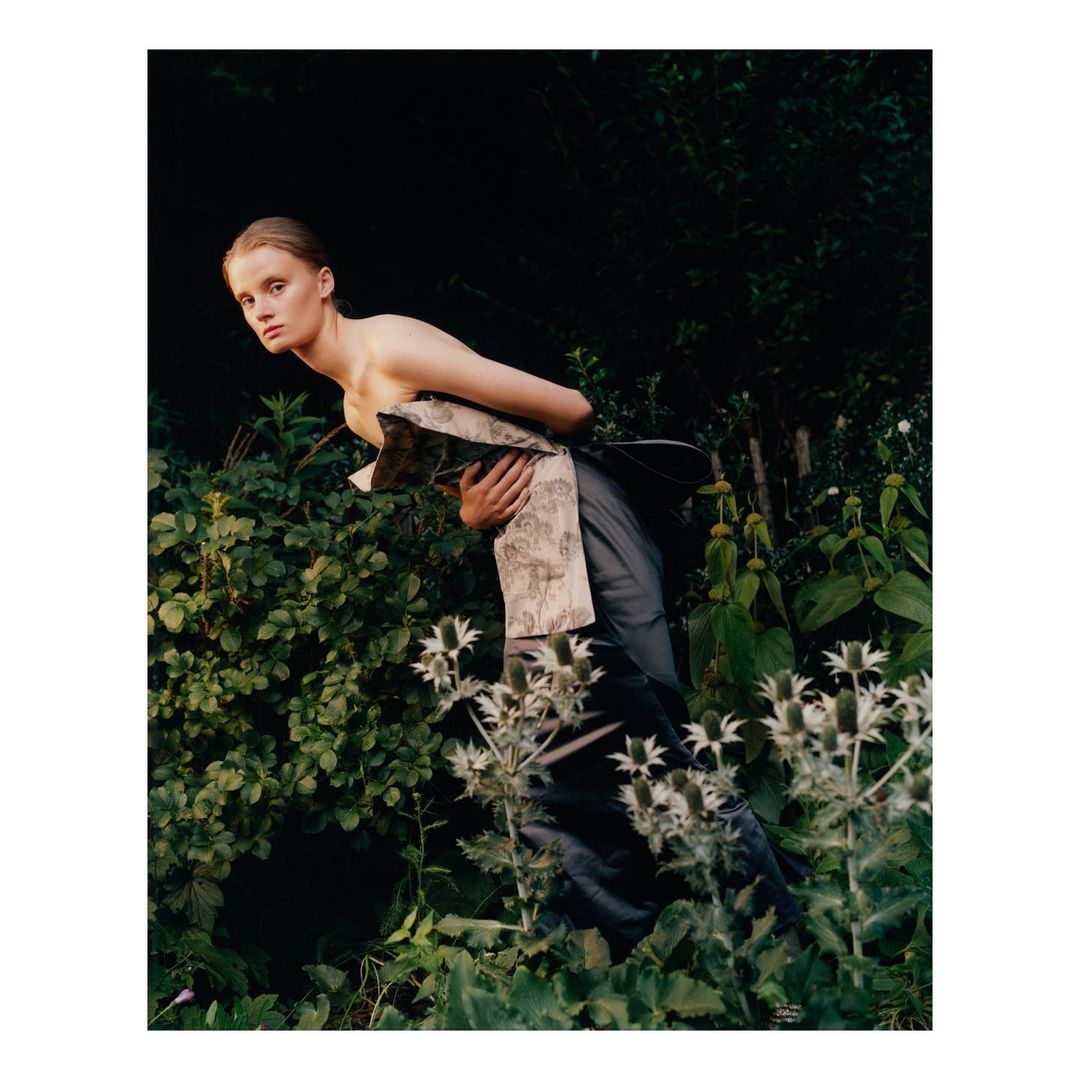 A fair-skinned adult woman poses in an overgrown garden wearing a dark sleeveless gown and a patterned shawl, with a neutral expression.