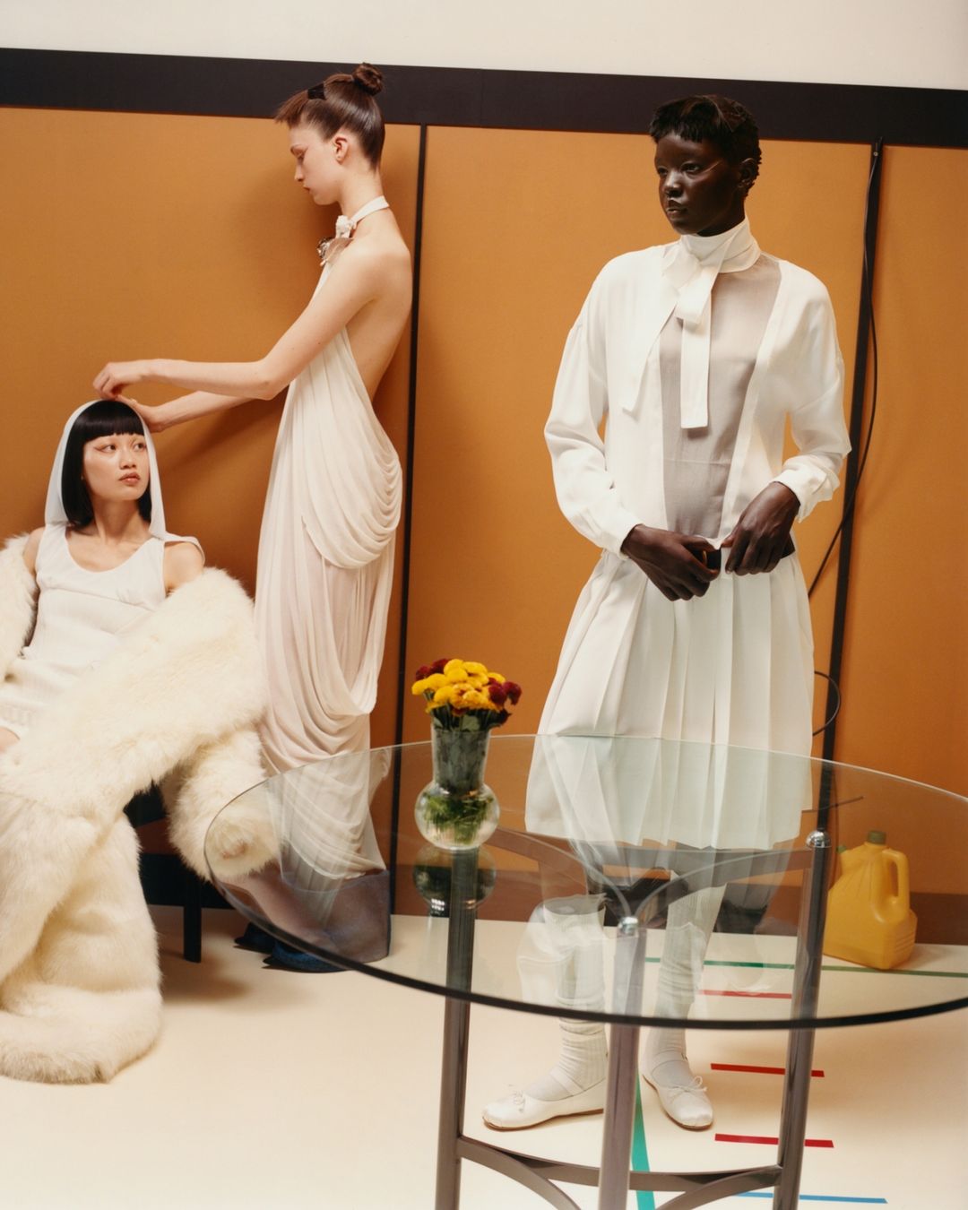 Three fashion models in an indoor setting. One is standing in a white dress, another is seated, and a third adjusts the seated model's hair.
