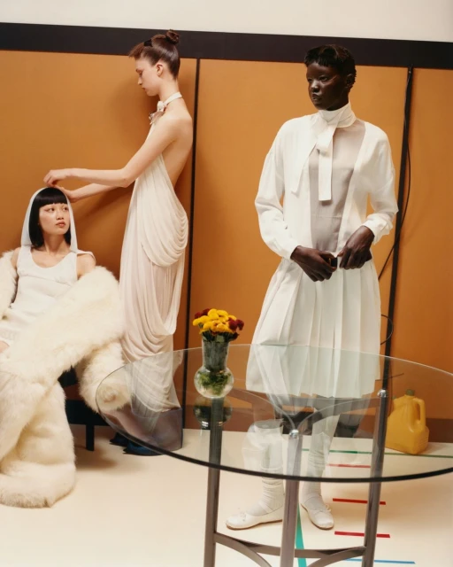 Three fashion models in an indoor setting. One is standing in a white dress, another is seated, and a third adjusts the seated model's hair.