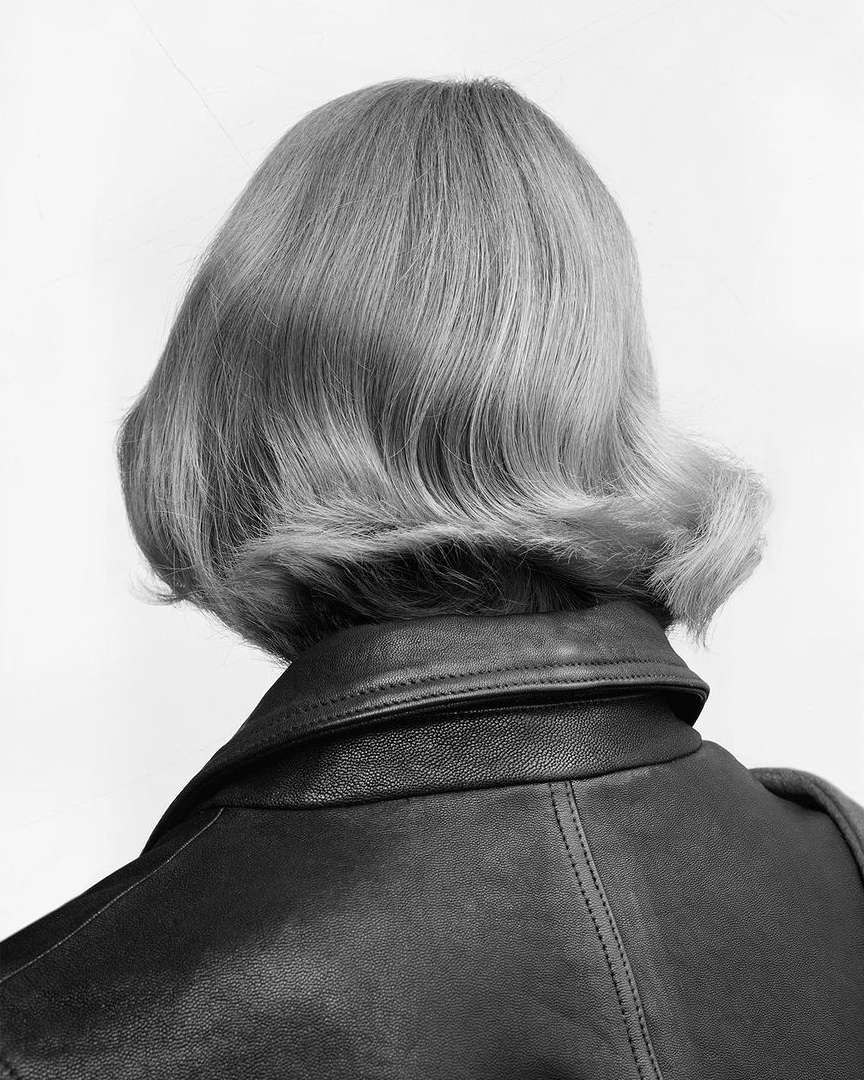 Close-up of the back of an adult female's head with a short, curled, blonde hairstyle and wearing a black leather jacket against a white background.