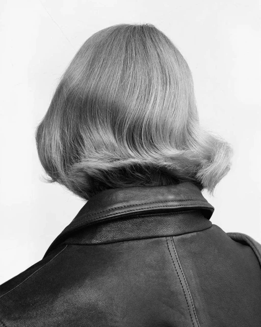 Close-up of the back of an adult female's head with a short, curled, blonde hairstyle and wearing a black leather jacket against a white background.