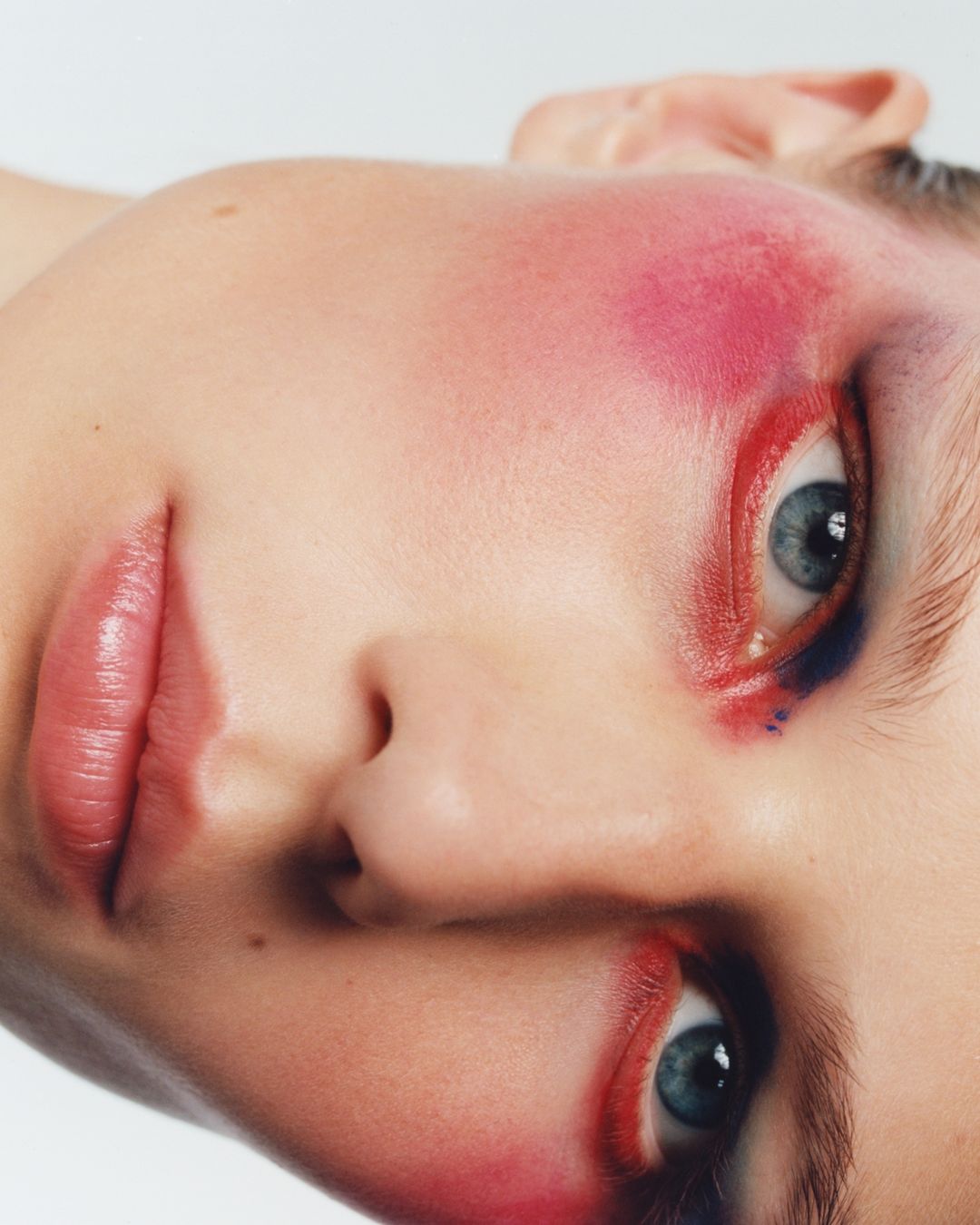 Close-up portrait of a woman's face with vibrant red and pink makeup around her eyes and cheeks against a plain white background.