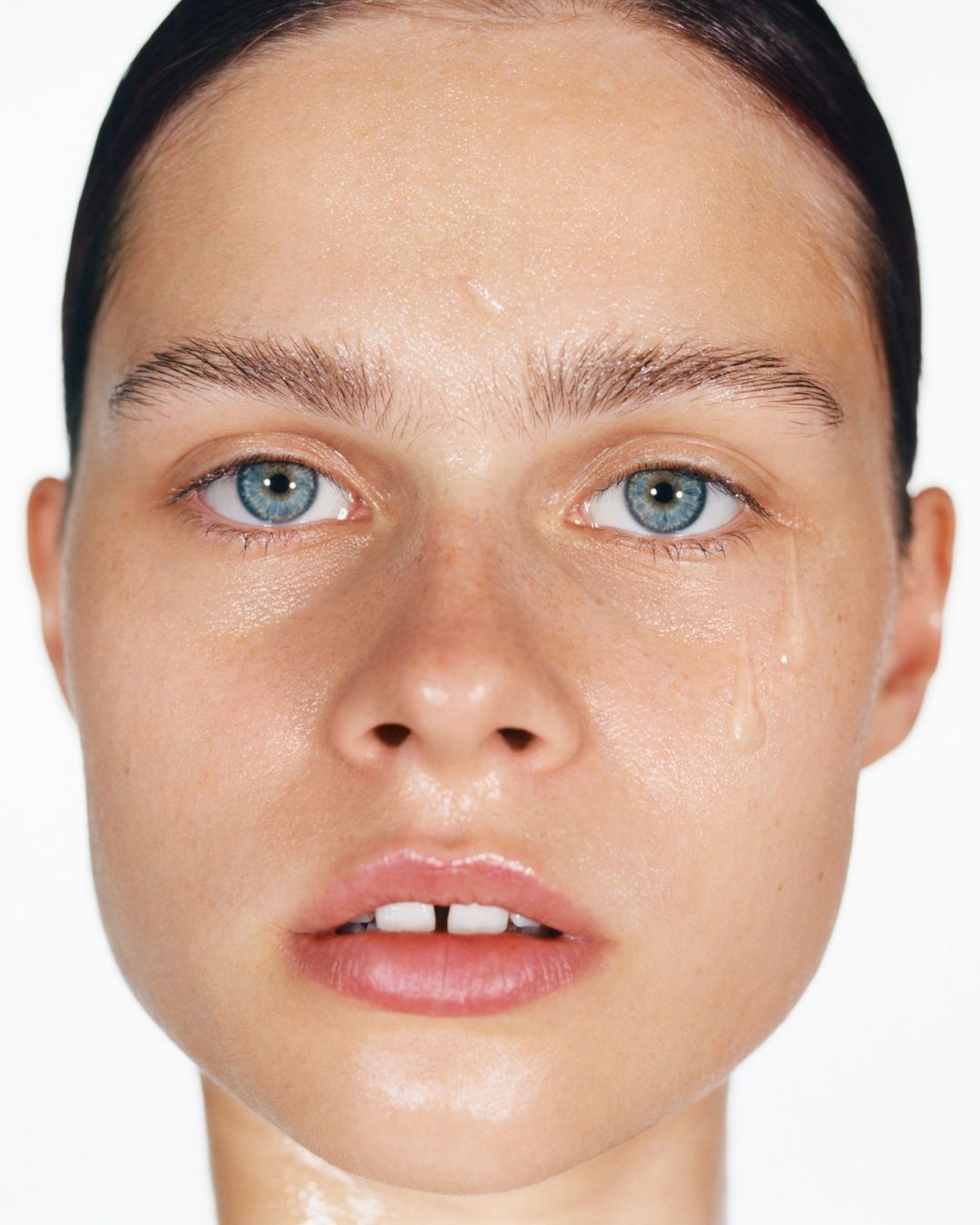 Close-up portrait of a young adult woman with blue eyes and fair skin with a visible tear, against a stark white background.