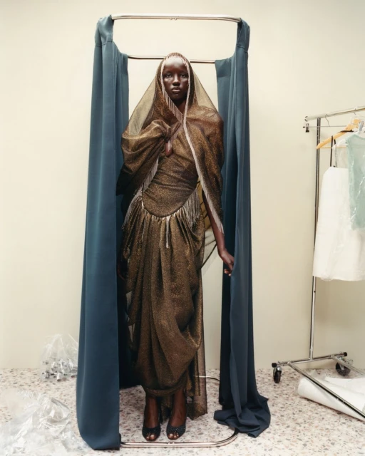 Woman in elegant gold gown with head covering stands in front of blue curtains and clothing rack, set against a neutral backdrop.