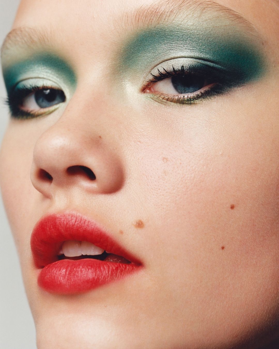 Close-up portrait of an adult female's face featuring prominent green eyeshadow, red lipstick, and clear skin with distinct beauty marks.